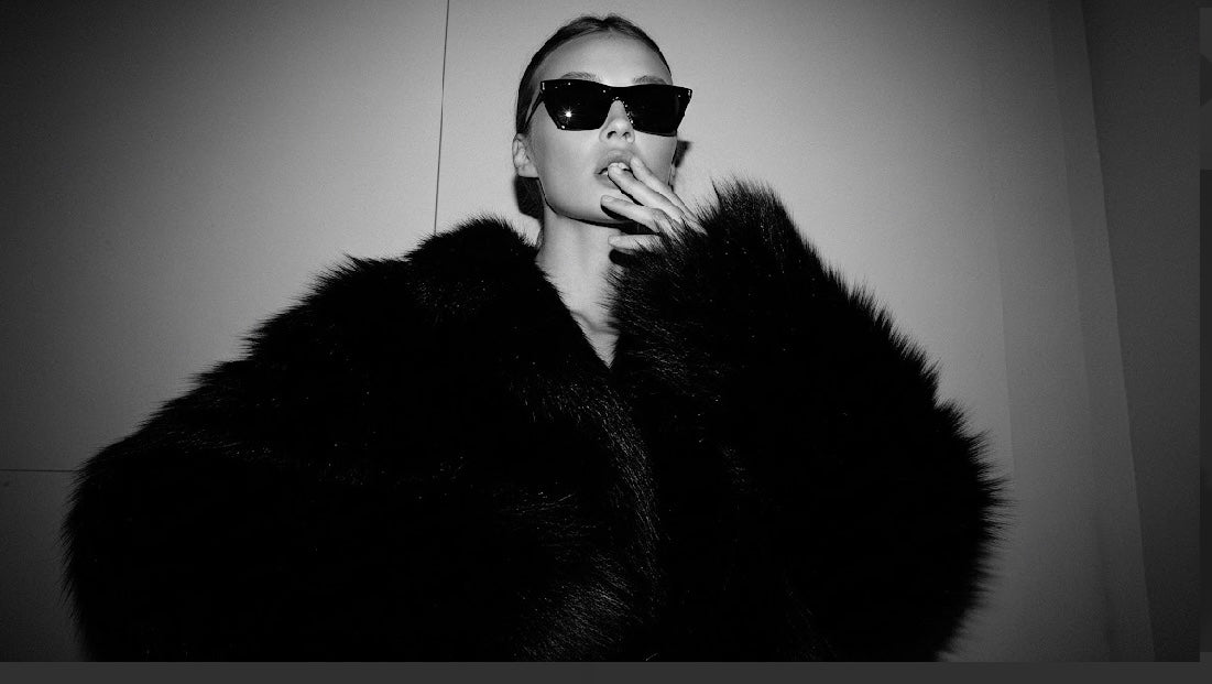  A person wearing oversized sunglasses and a dark blazer poses playfully with their tongue out, touching their chin with one hand in a high-contrast black-and-white portrait.