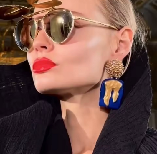 Person wearing gold aviator sunglasses and blue earrings with a blurred background